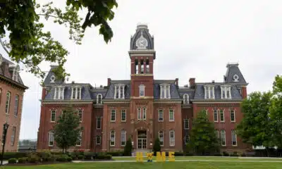 A red brick building with a clock tower in the center sit behind a gold sign that says "home" with the "o" replaced with West Virginia University's Flying WV logo.